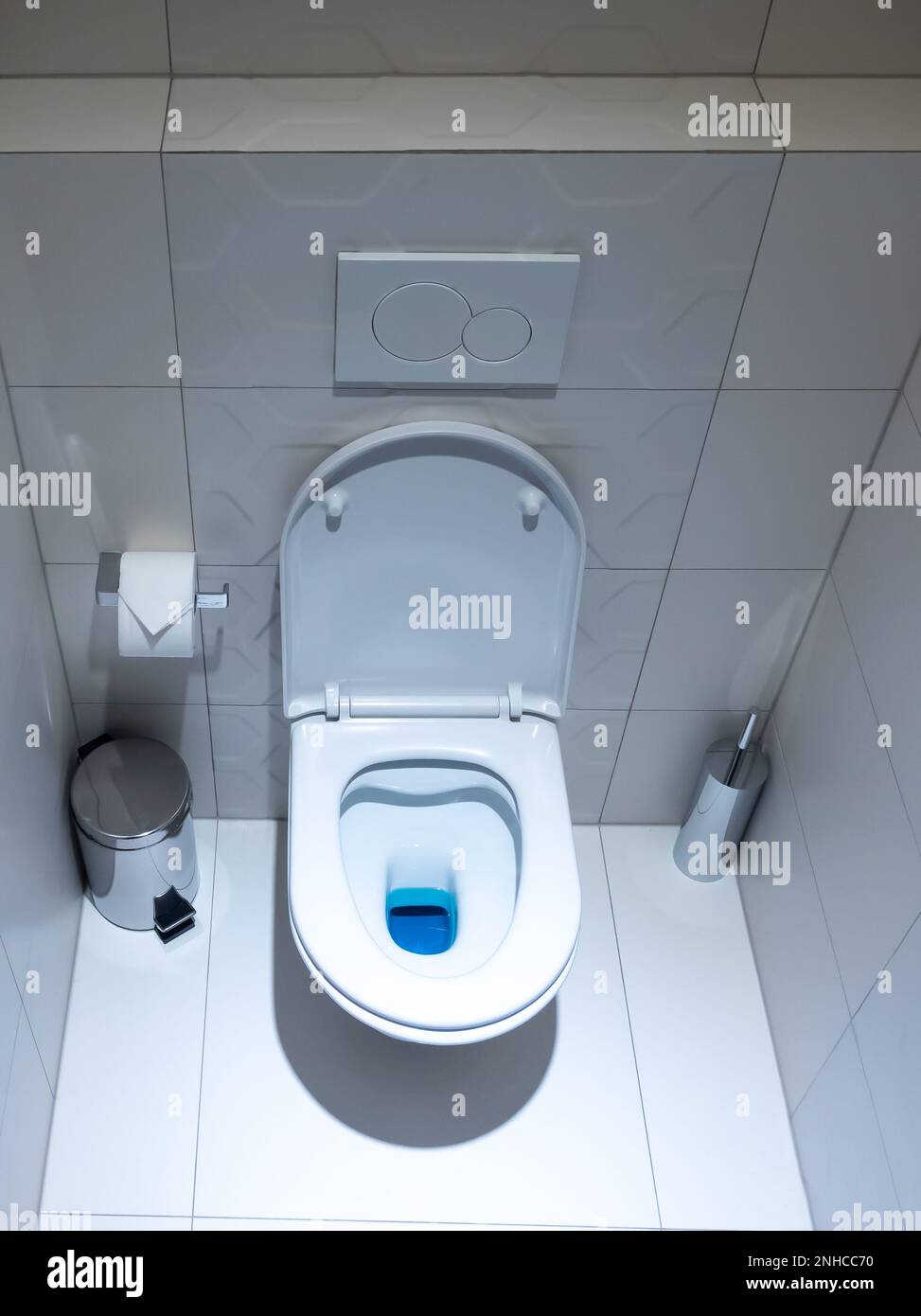 View of the white toilet bowl in the toilet. Bathroom finished with white glaze tiles and white terracotta. Trash can and brush in the background Stock Photo