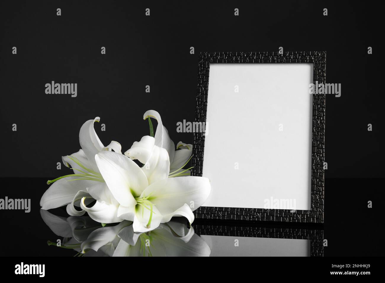 Funeral photo frame and white lilies on black table against dark