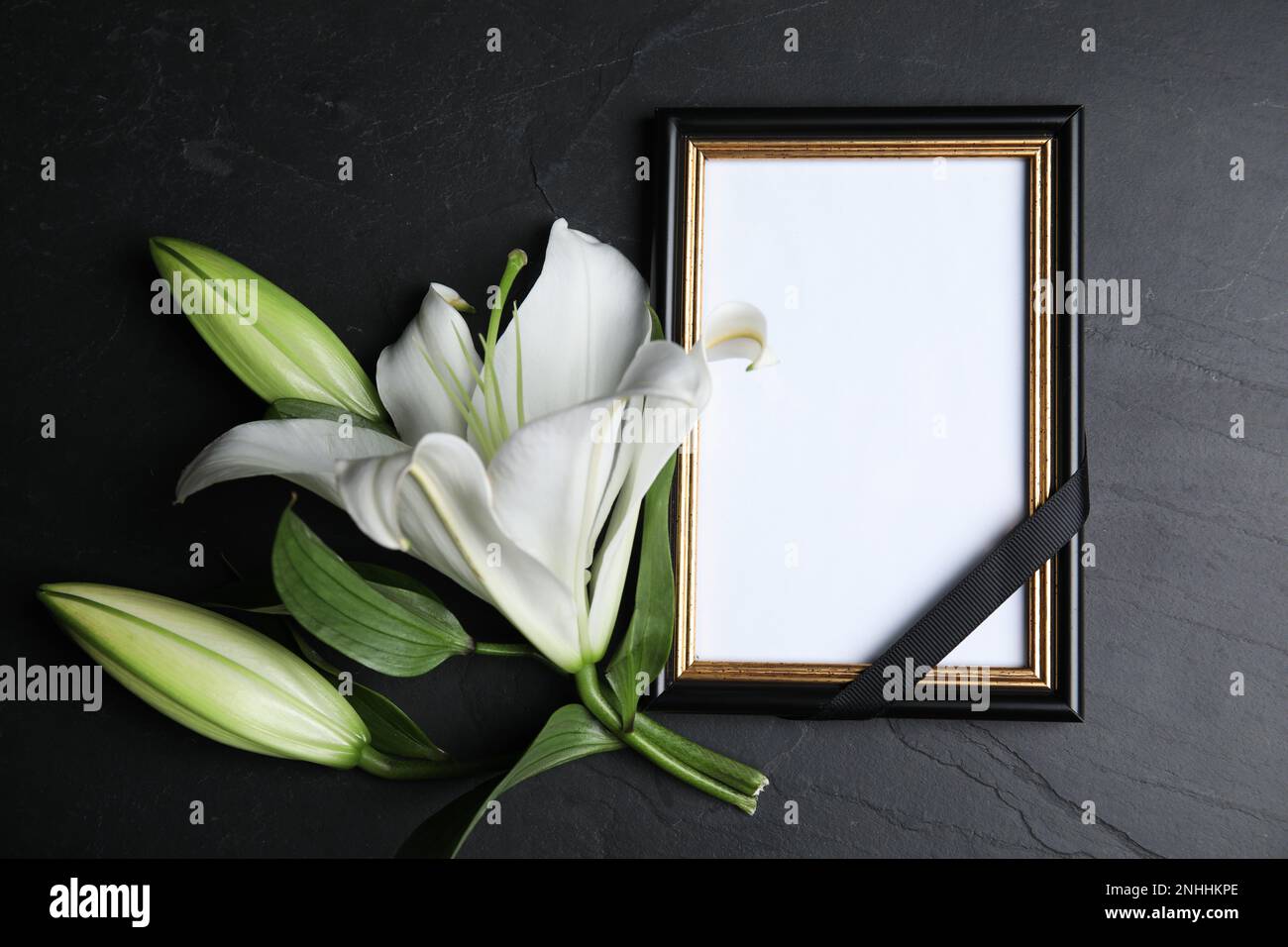 Funeral photo frame with ribbon and white lily on black table, flat lay