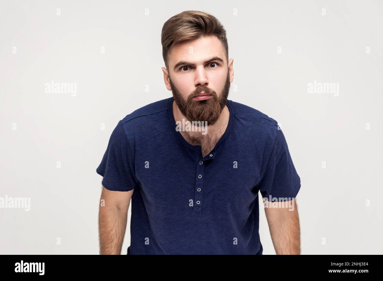Portrait of disgruntled man with beard wearing dark blue T-shirt looking with annoyed indignant expression at camera, being angry and aggressive. Indoor shot isolated on gray background. Stock Photo