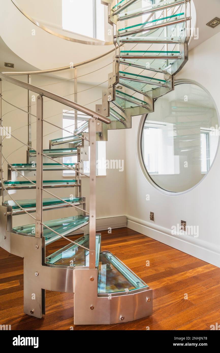 Steel and tempered glass spiraling staircase and wall with porthole window, exotic wood plank floor in hallway of master bedroom inside modern condo. Stock Photo