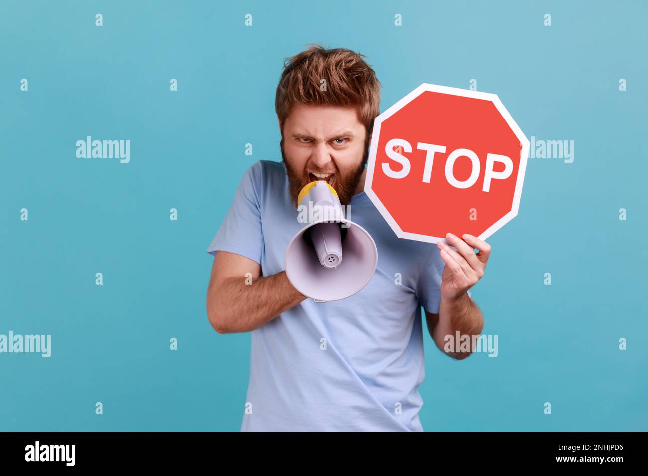 Portrait of aggressive angry bearded man looking at camera, screaming in loud speaker, holding red stop sign and megaphone in his hands. Indoor studio shot isolated on blue background. Stock Photo