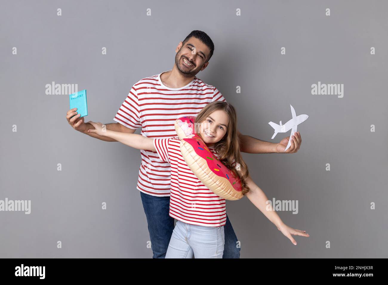 Portrait of excited smiling father and daughter in striped T-shirts holding passport and rubber ring, journey abroad, vacation to the sea. Indoor studio shot isolated on gray background. Stock Photo