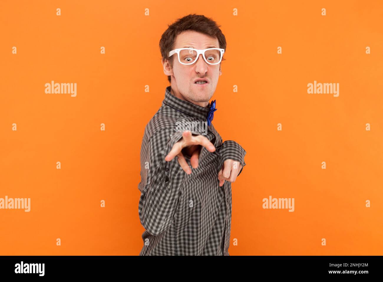 Portrait of funny crazy disabled man nerd pointing and looking at camera with clenched teeth, wearing shirt with blue bow tie and white glasses. Indoor studio shot isolated on orange background. Stock Photo