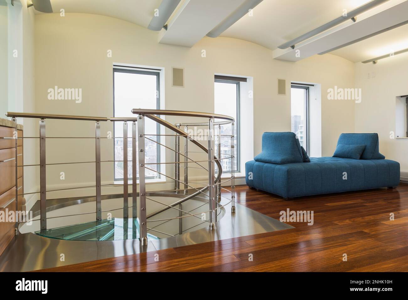 Steel and tempered glass spiraling staircase with blue cloth upholstered daybed in home office with exotic wood cabinet and floor inside condo unit. Stock Photo