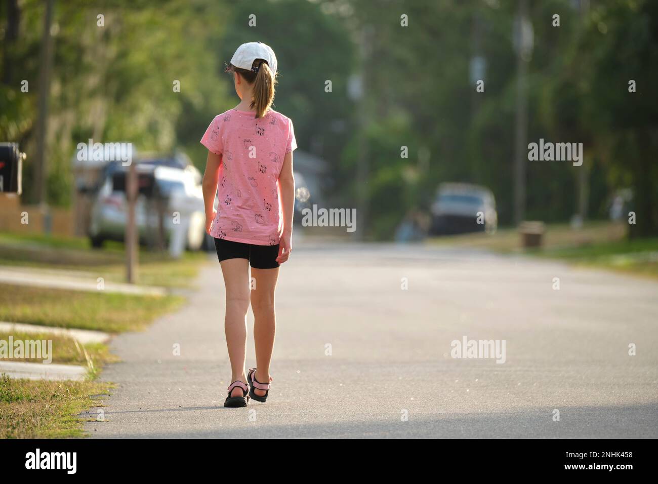 Rear view of confident young child girl walking along the sunny alley ...