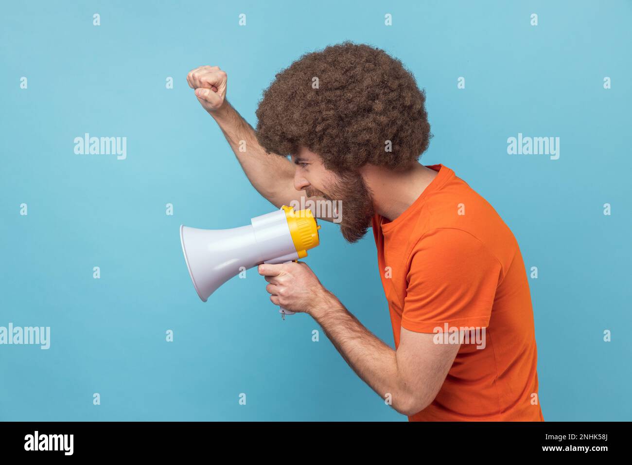 Side view of serious man with Afro hairstyle wearing orange T-shirt raised hands and holding megaphone, screaming in loud speaker, protesting. Indoor studio shot isolated on blue background. Stock Photo