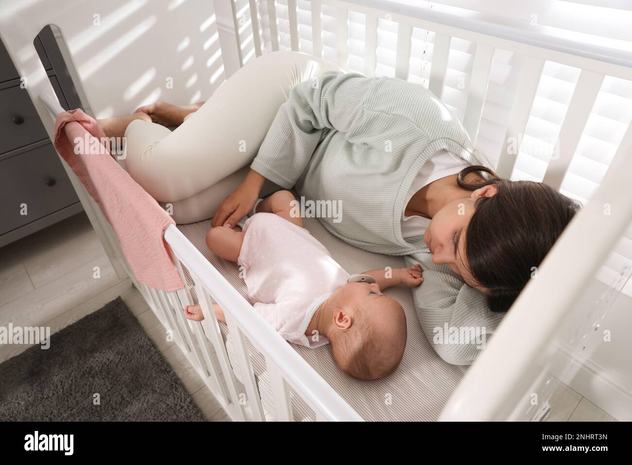 Tired young mother sleeping in crib with her baby at home, above view