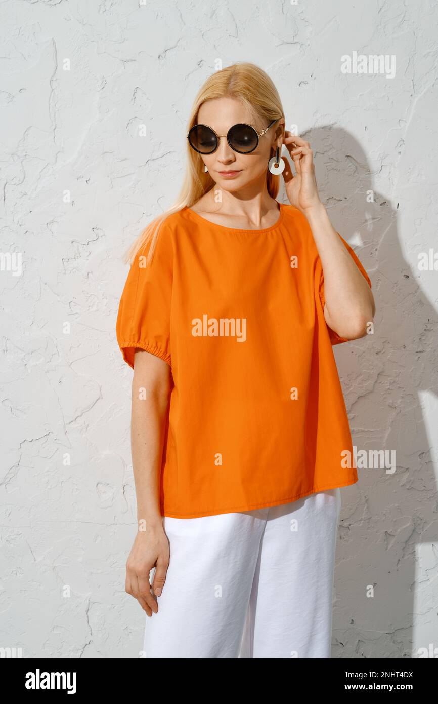 A fashionable woman stands in three-quarter length, wearing an orange top with short sleeves and sunglasses Stock Photo