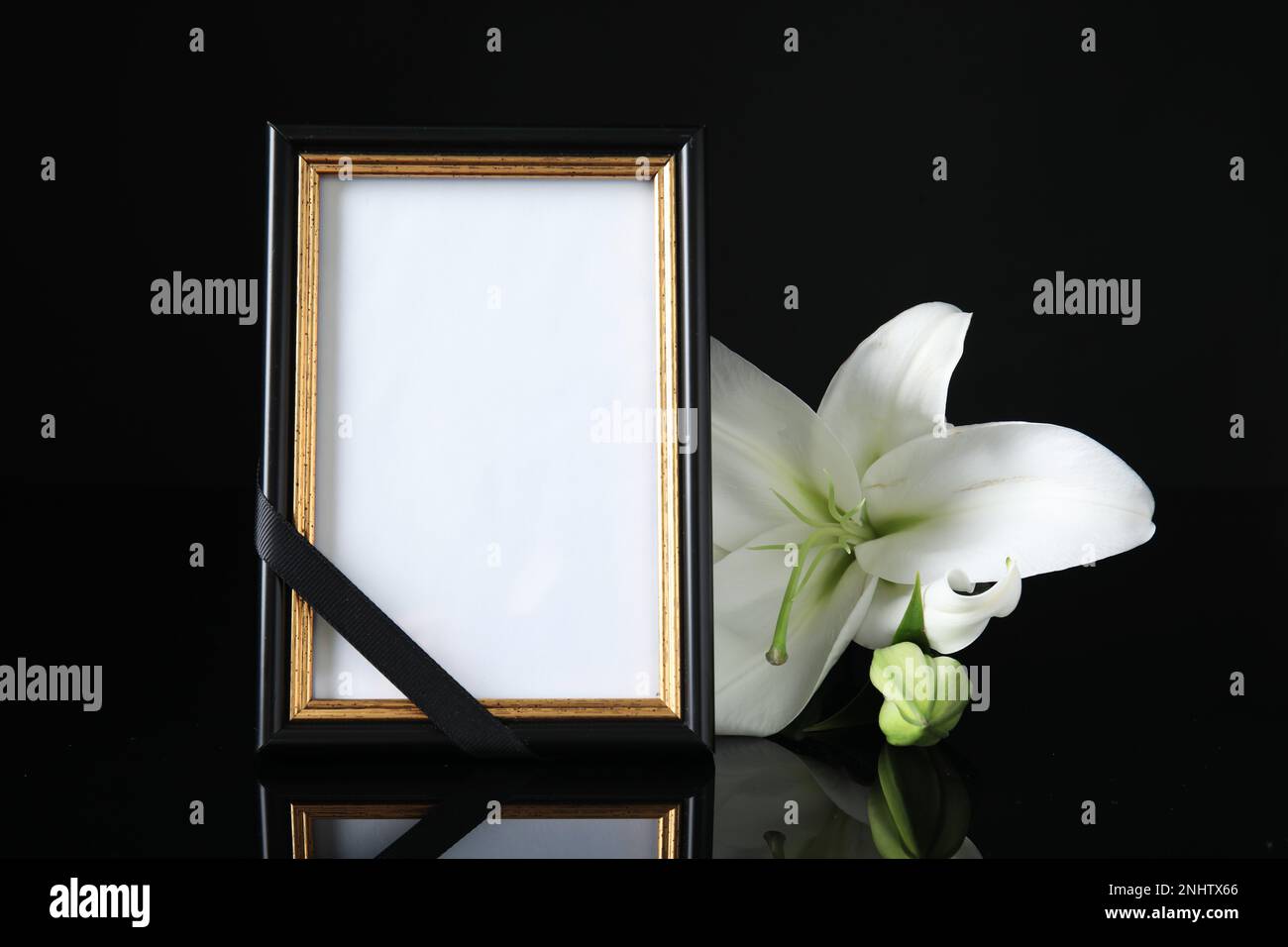 Funeral photo frame with ribbon and white lily on black table against dark background. Space for