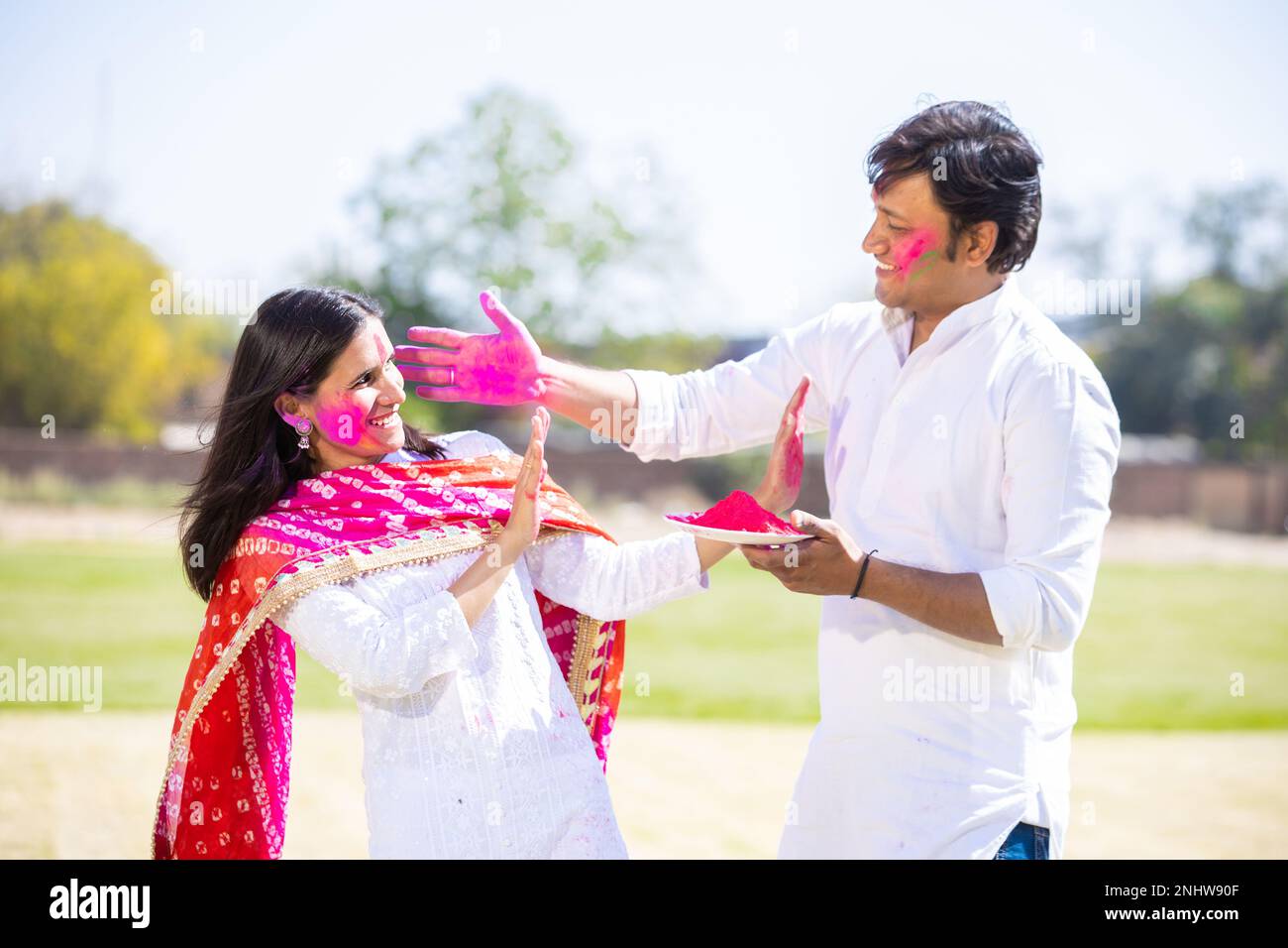 Happy young indian couple wearing white kurta playing celebrating holi together outdoor at park. Stock Photo