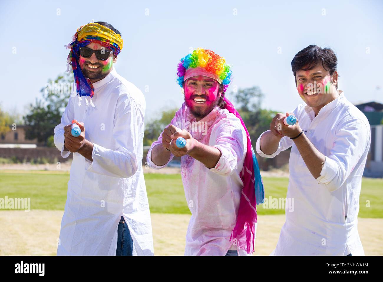 Cheerful young indian male friends wearing white kurta outfit playing with pichkari celebrating holi festival outdoor at park. Stock Photo
