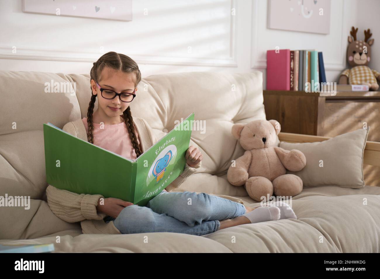 Cute little girl reading book on sofa at home Stock Photo - Alamy