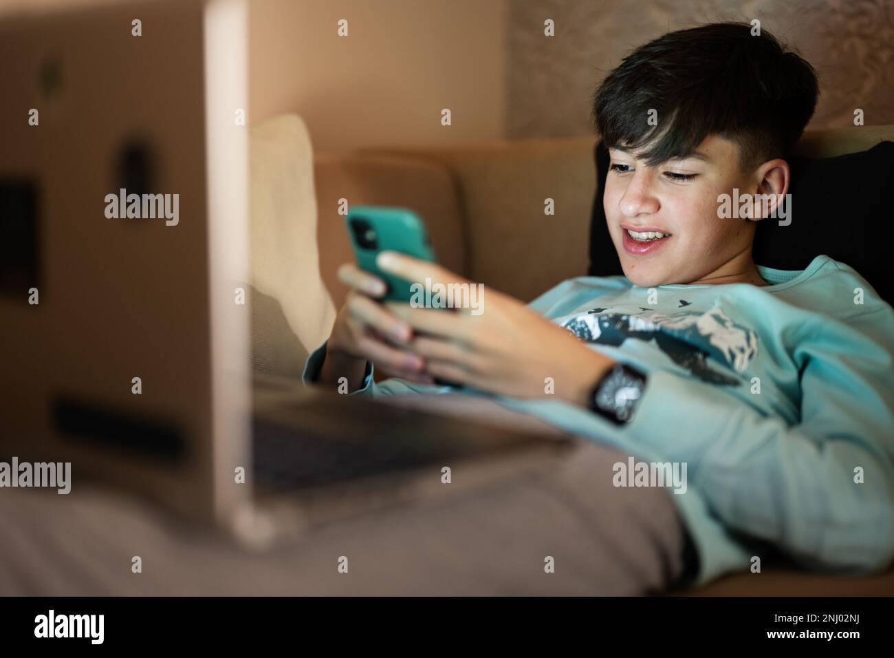 Young teen boy texting phone in front of a laptop on a bed at evening. Stock Photo