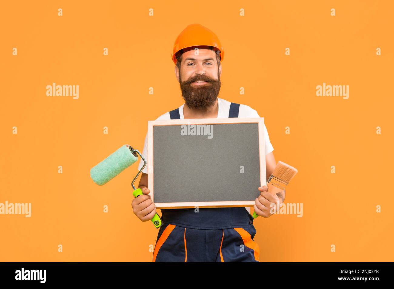 happy man renovator in helmet and work clothes hold paint roller brush