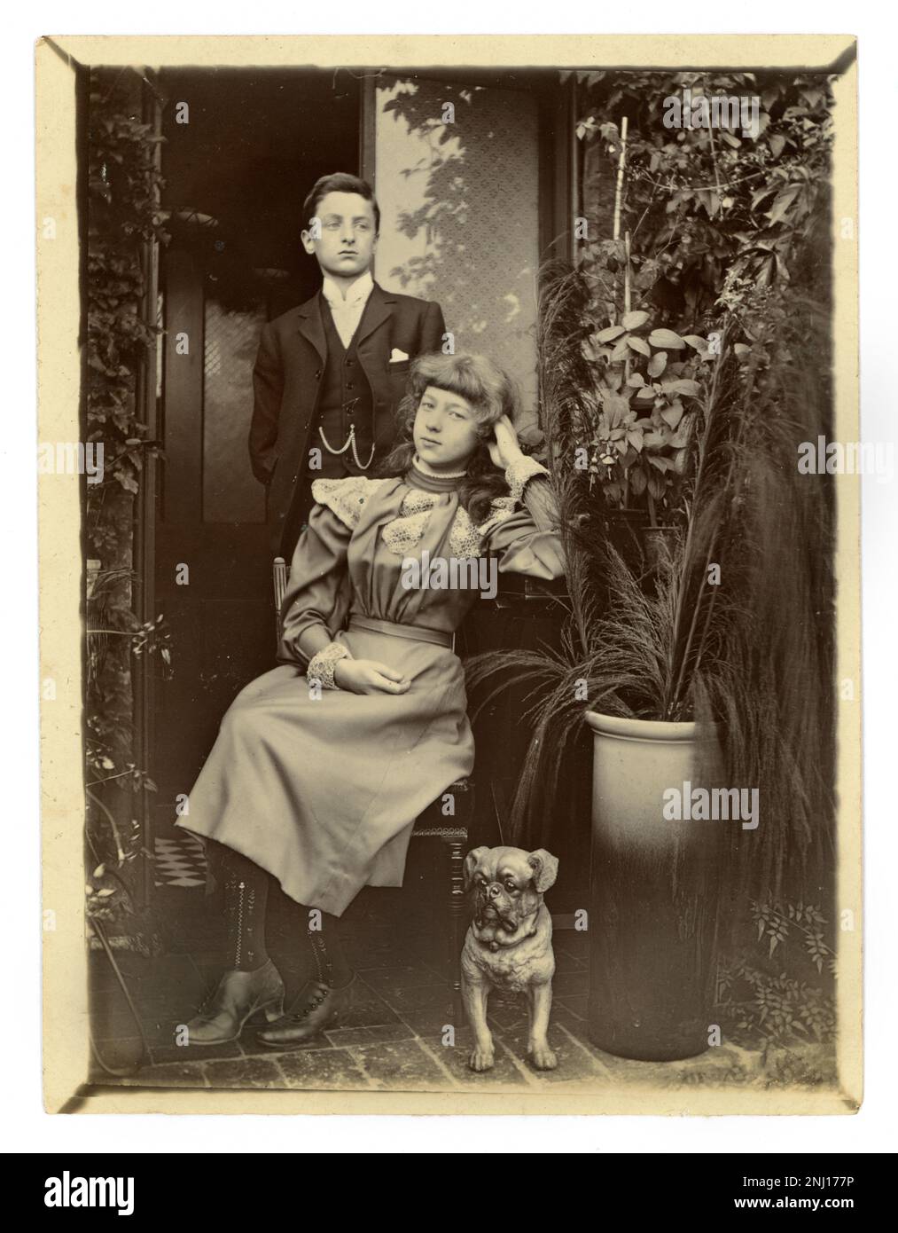 Original Victorian era photograph of stroppy / belligerent looking teenagers - a middle class attractive boy and girl, probably brother and sister. A small pug dog sits next to them, wearing a collar with bells on it - a naughty one! It's a relaxed, informal photograph taken outside a house in the doorway of their home, potted plants next to them, circa 1899 Stock Photo