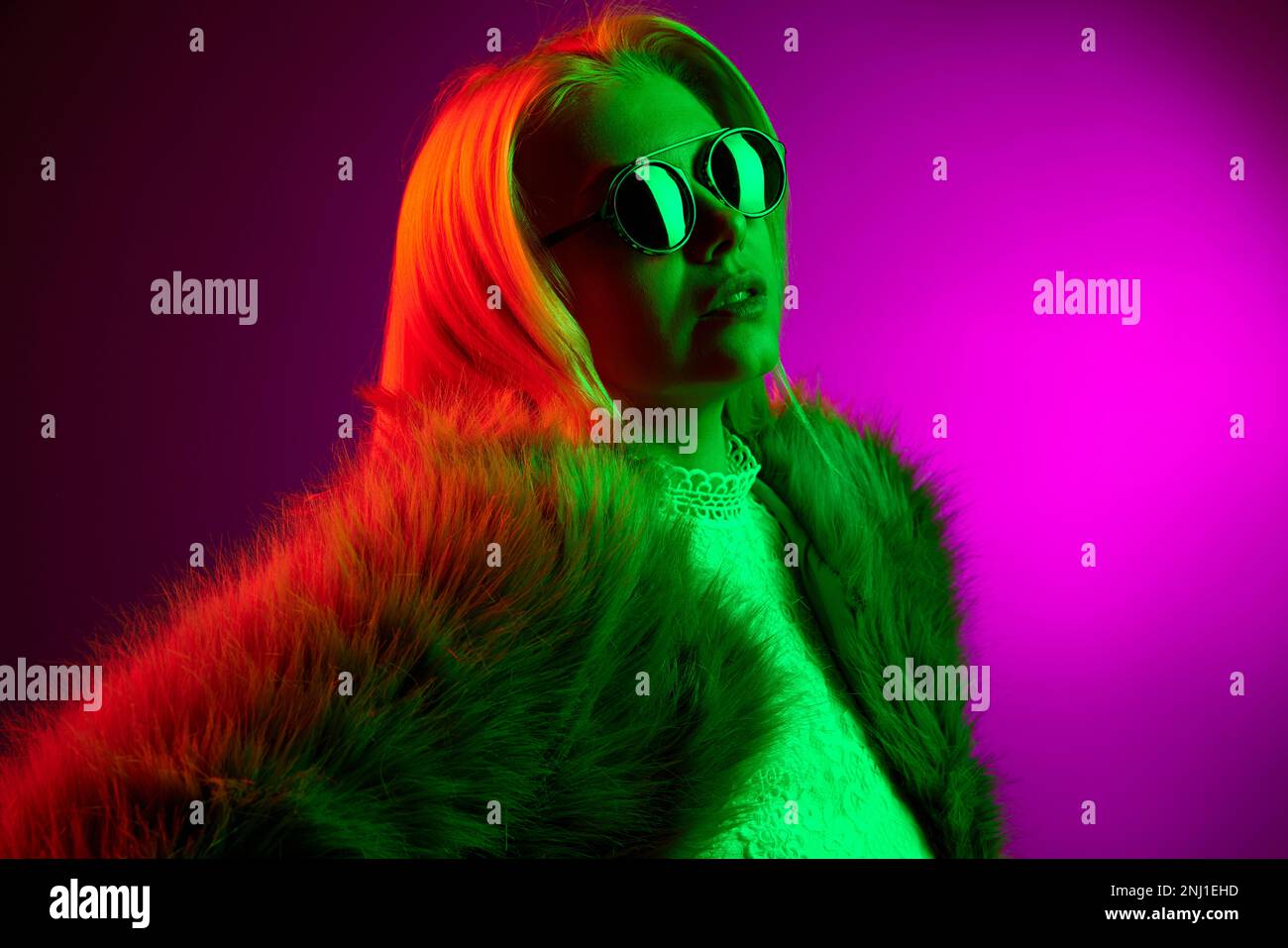 Stylish confident young woman posing in fur coat and sunglasses over magenta studio background in green neon light. Concept of emotions, fashion, faci Stock Photo