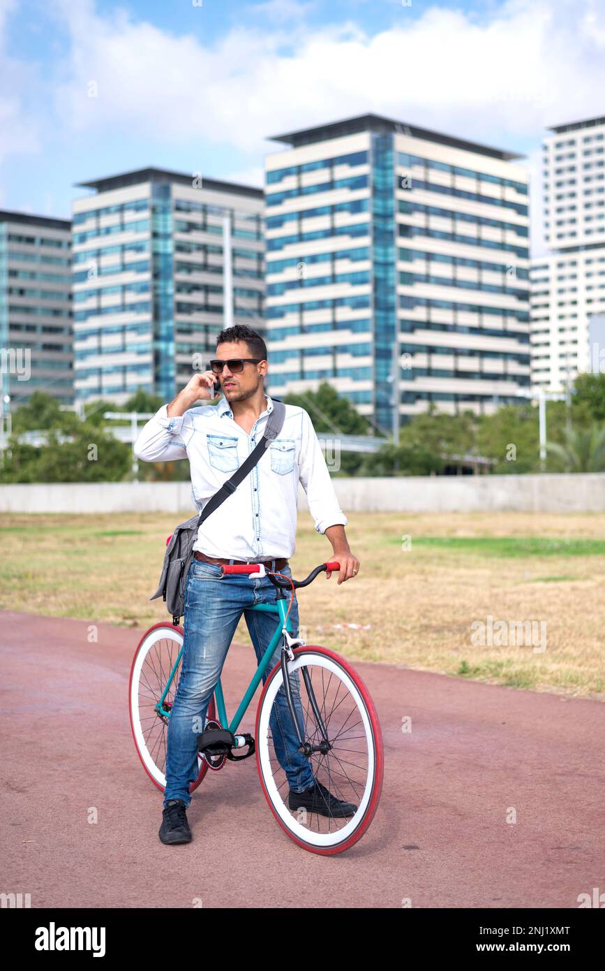A Man with sunglasses standing with his bicycle talking by phone and the city in the background Stock Photo