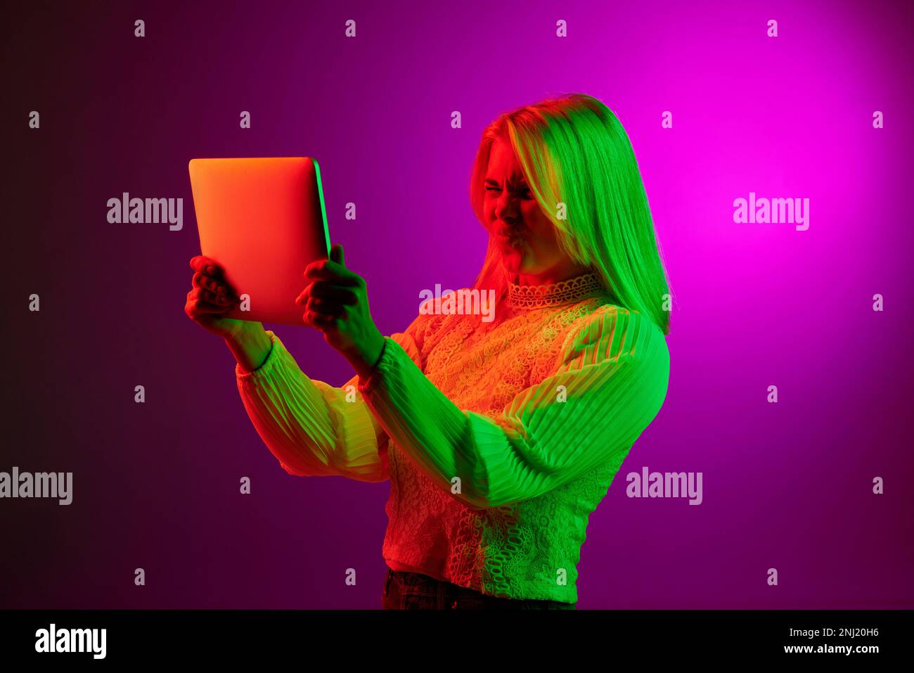 Communication. Emotive young woman having online video call on tablet over magenta studio background in green neon light. Concept of emotions, facial Stock Photo