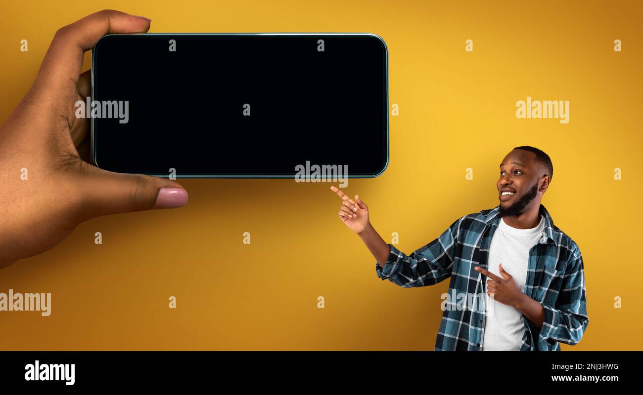 Positive Black Man Pointing Aside At Huge Female Hand With Blank Smartphone Stock Photo