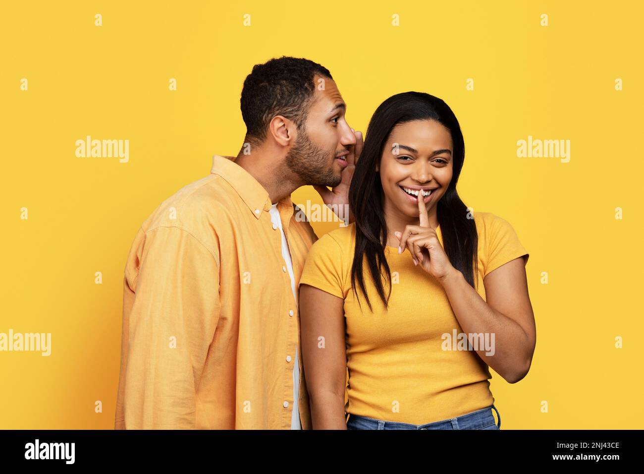 Smiling young african american male whispers to lady ear, female puts finger to lips, making secret gesture Stock Photo