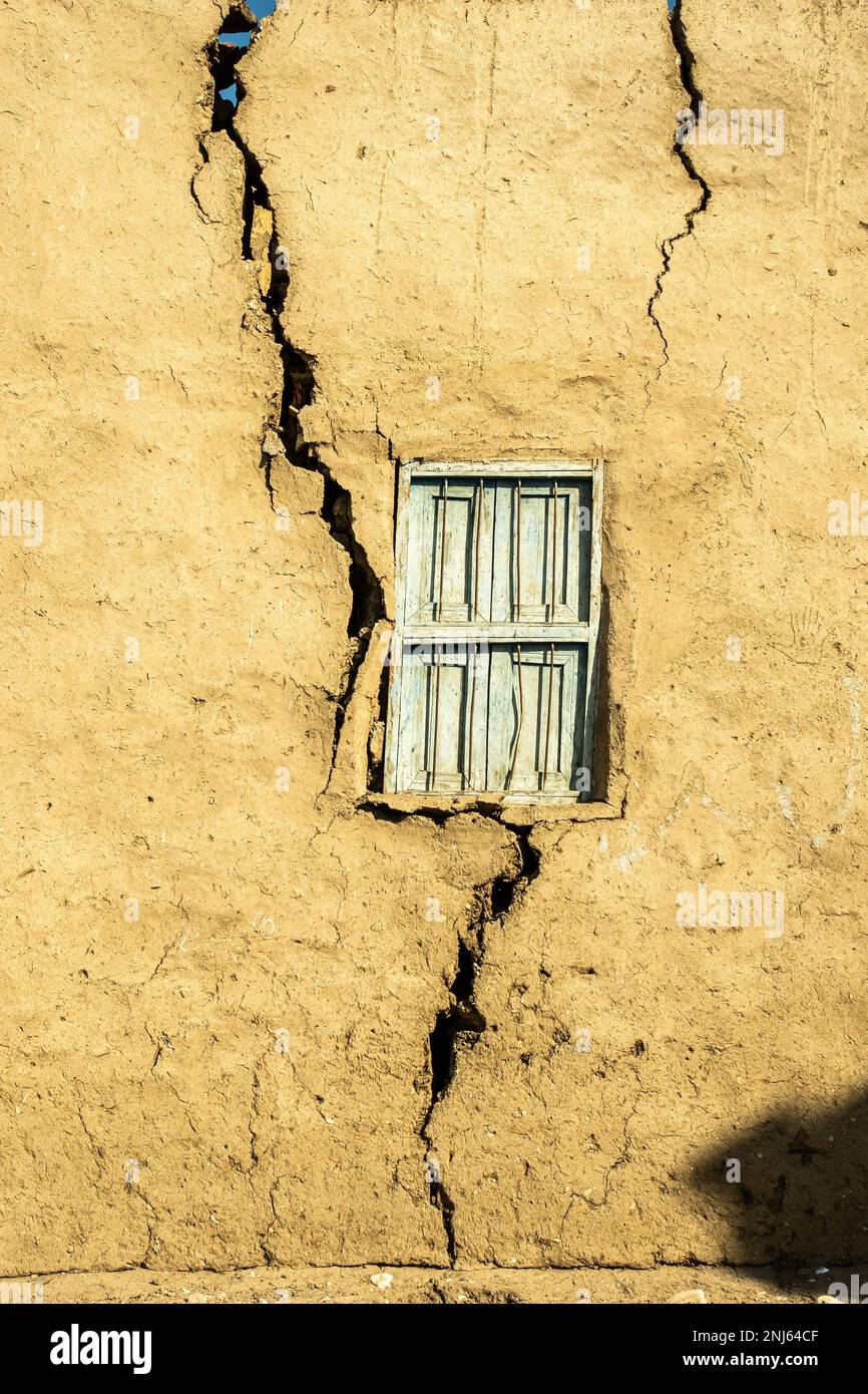 clay wall with crack. blue wooden window in the middle of the wall. traditional construction with clay and straw. Stock Photo