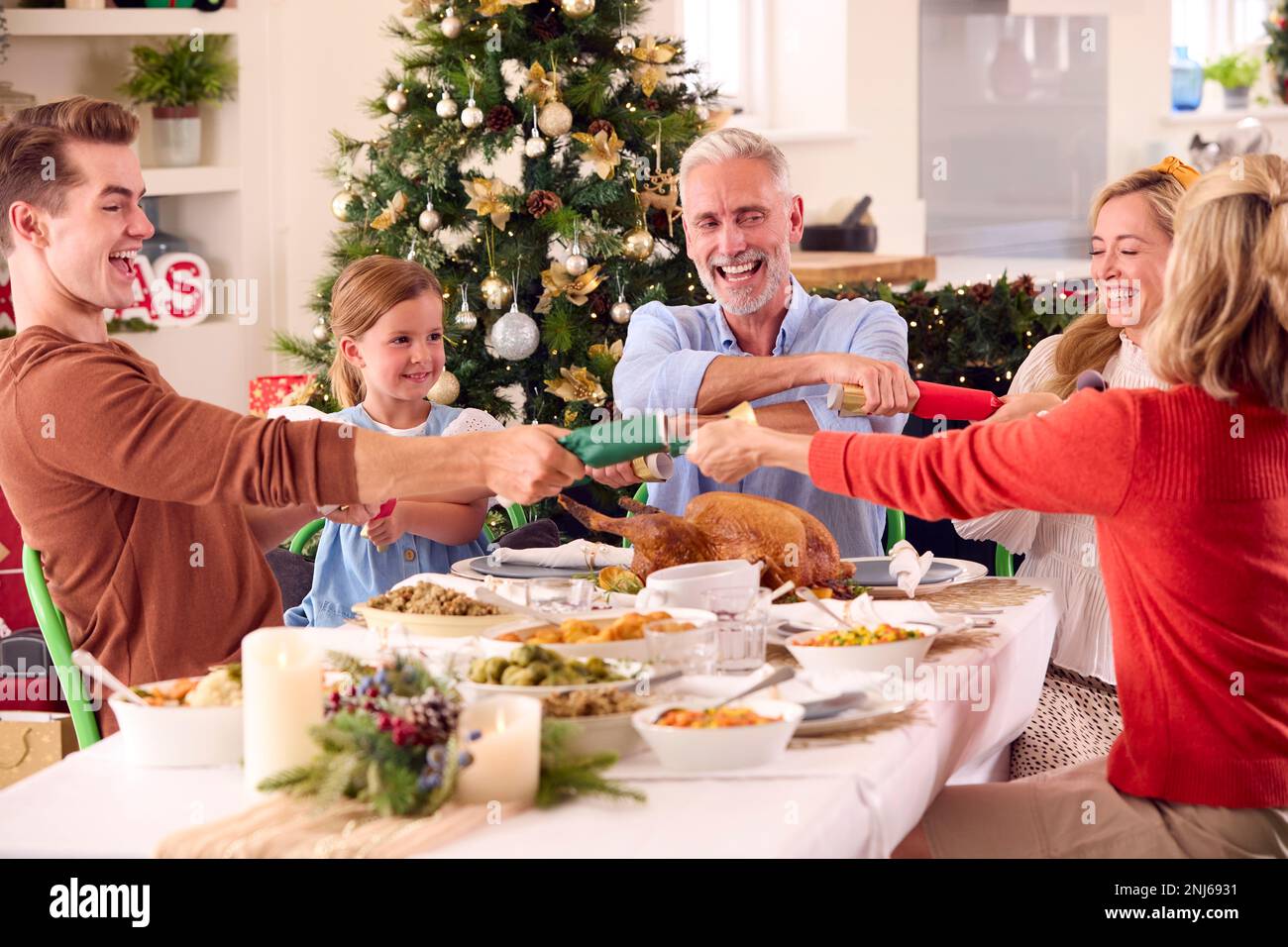 Multi-Generation Family Celebrating Christmas At Home Pulling Crackers Before Eating Meal Together Stock Photo