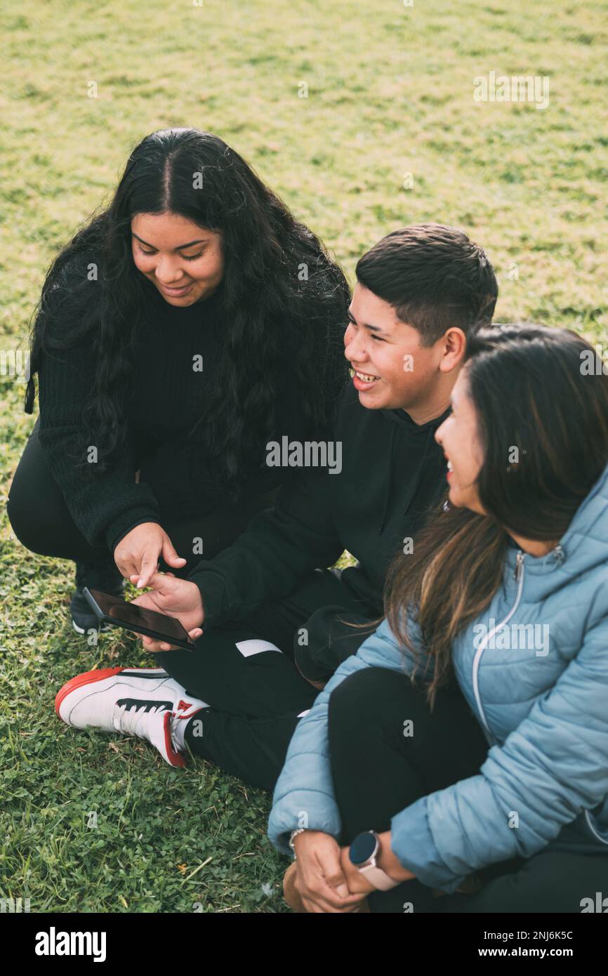 Hispanic male teenager holding smartphone looking away while sitting on grass with Hispanic mother and sister in park on sunny day Stock Photo