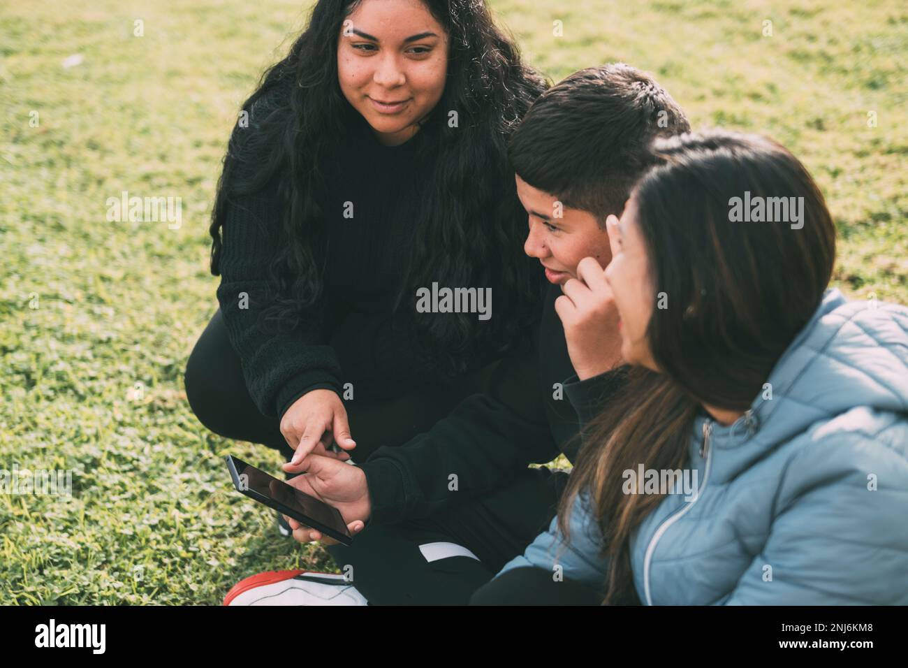 Hispanic family spending quality time together in a local park on a beautiful, sunny day. A teenage boy is sitting on the grass, holding his Stock Photo