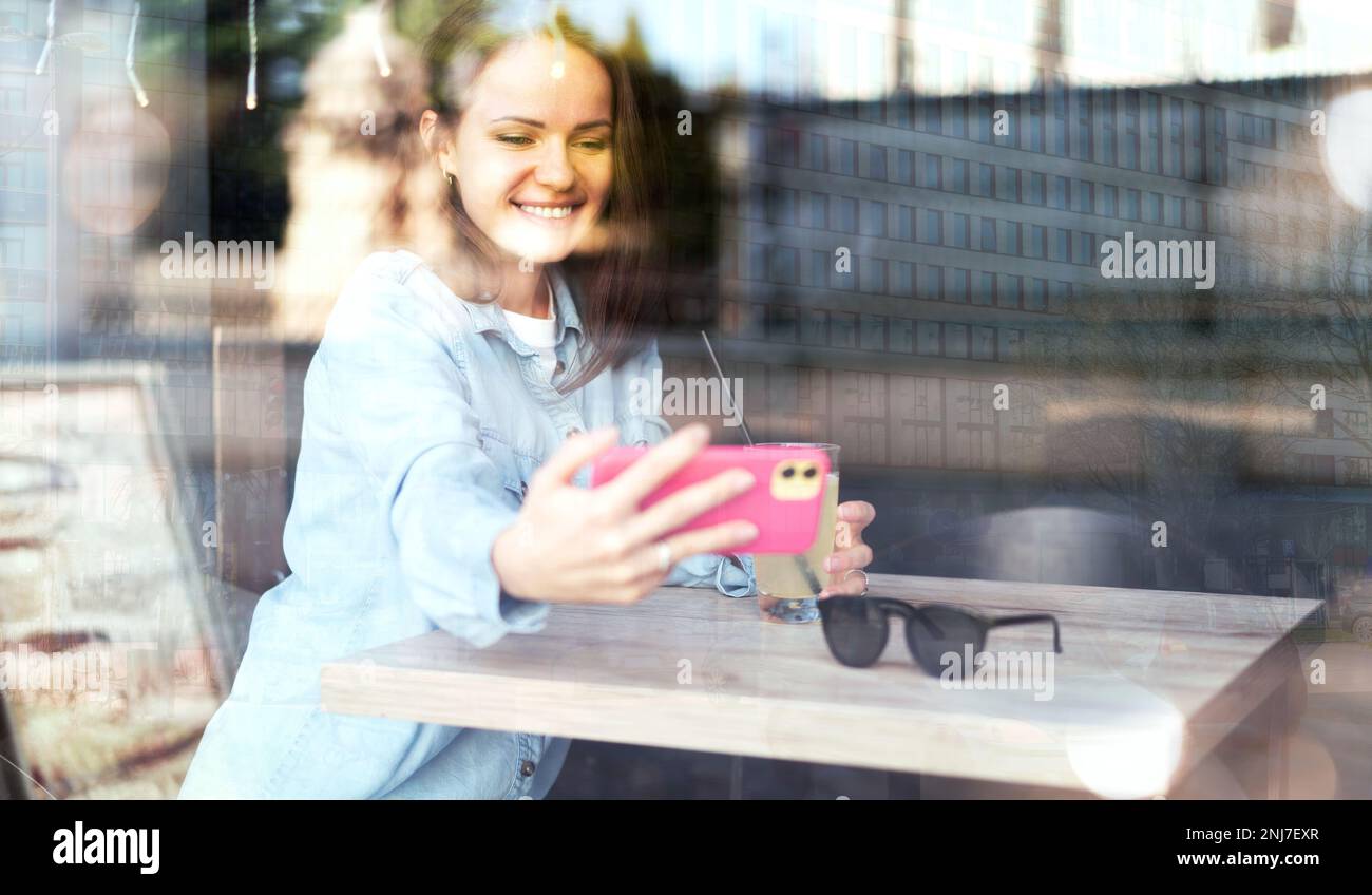 Woman sitting inside cafe taking a selfie using her smartphone, reflection of urban street on glass of window of cafe. Stock Photo