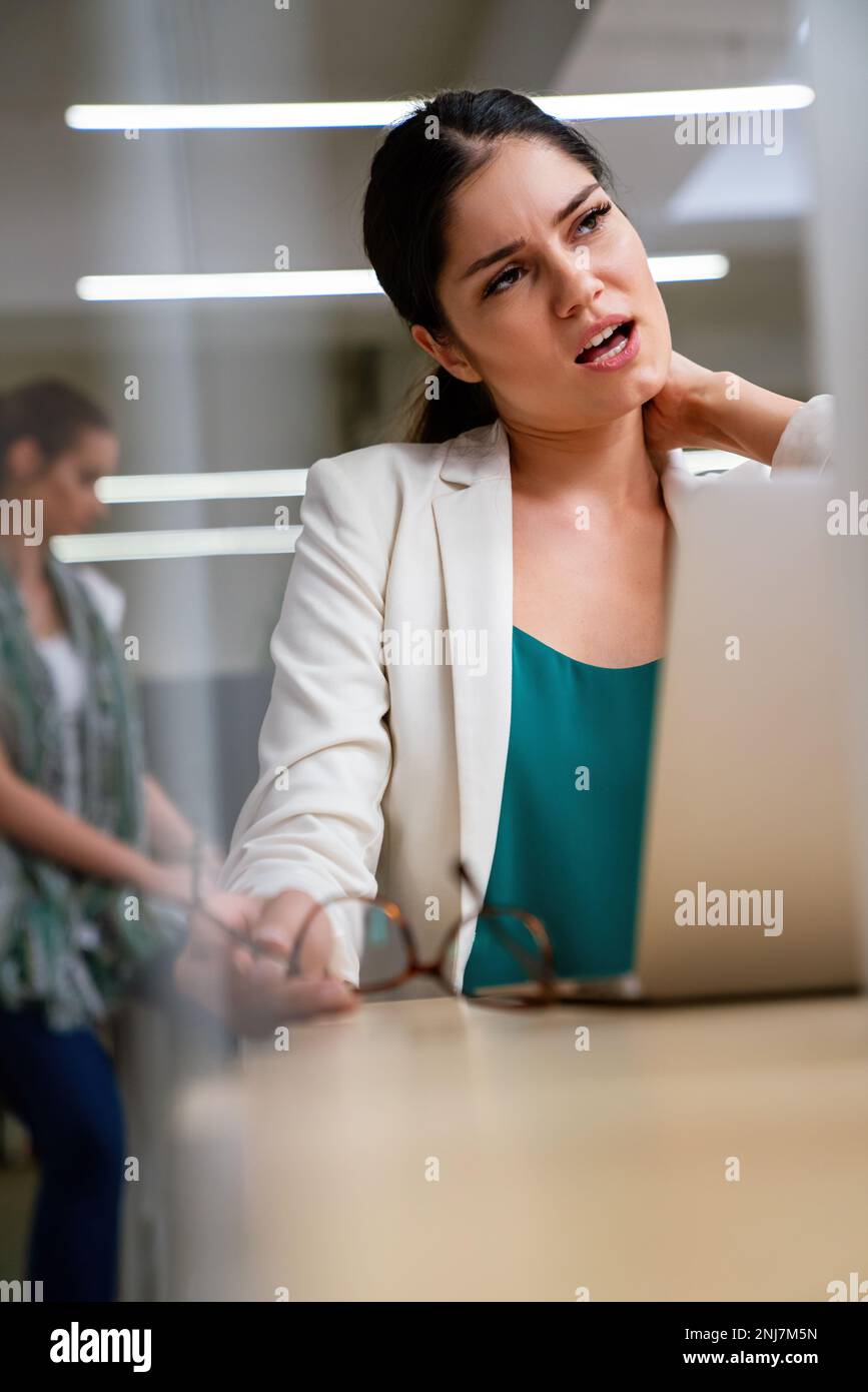 Young woman at office desk working on laptop with stress, neck pain and