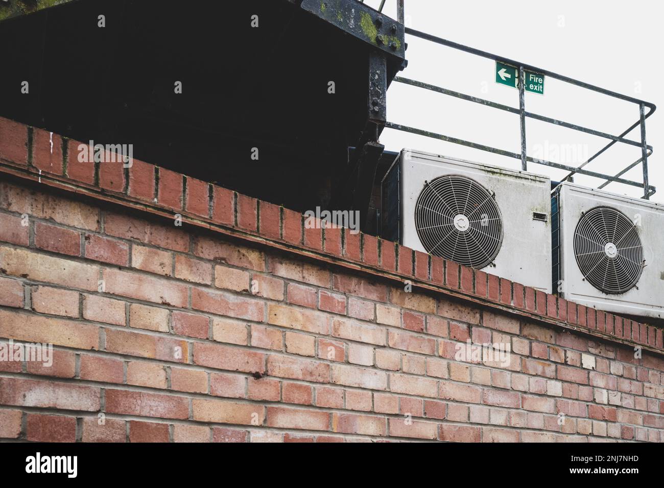 Pair of office air conditioning units seen perched atop a brick wall. A fire exit sign and gantry is seen directly above. Stock Photo