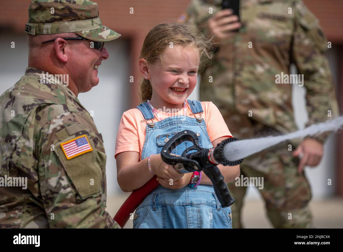 Six-year-old Birdie Amerson sprays a fire hose with the help of Sgt. Steve Krueger, 467th Engineer Battalion, Firefighter Headquarters, during an open house at Fort Des Moines, Iowa, Aug. 6, 2022. The event welcomed more than 70 people to the historic U.S Army Reserve base to see the current training units. Stock Photo