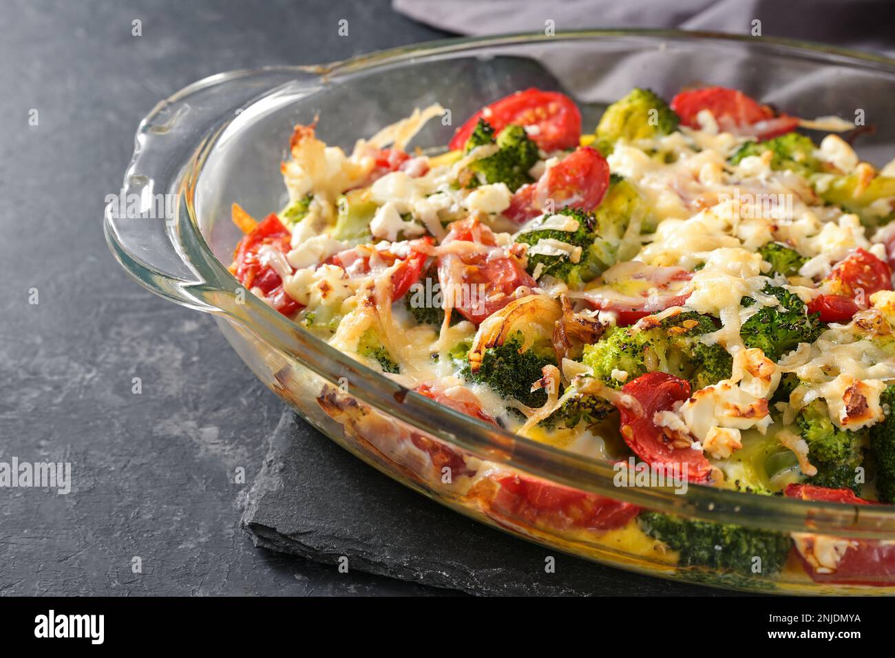 Vegetable dish from broccoli, tomatoes, onions, feta and cheese in a glass casserole on a gray slate table, copy space, selected focus, narrow depth o Stock Photo