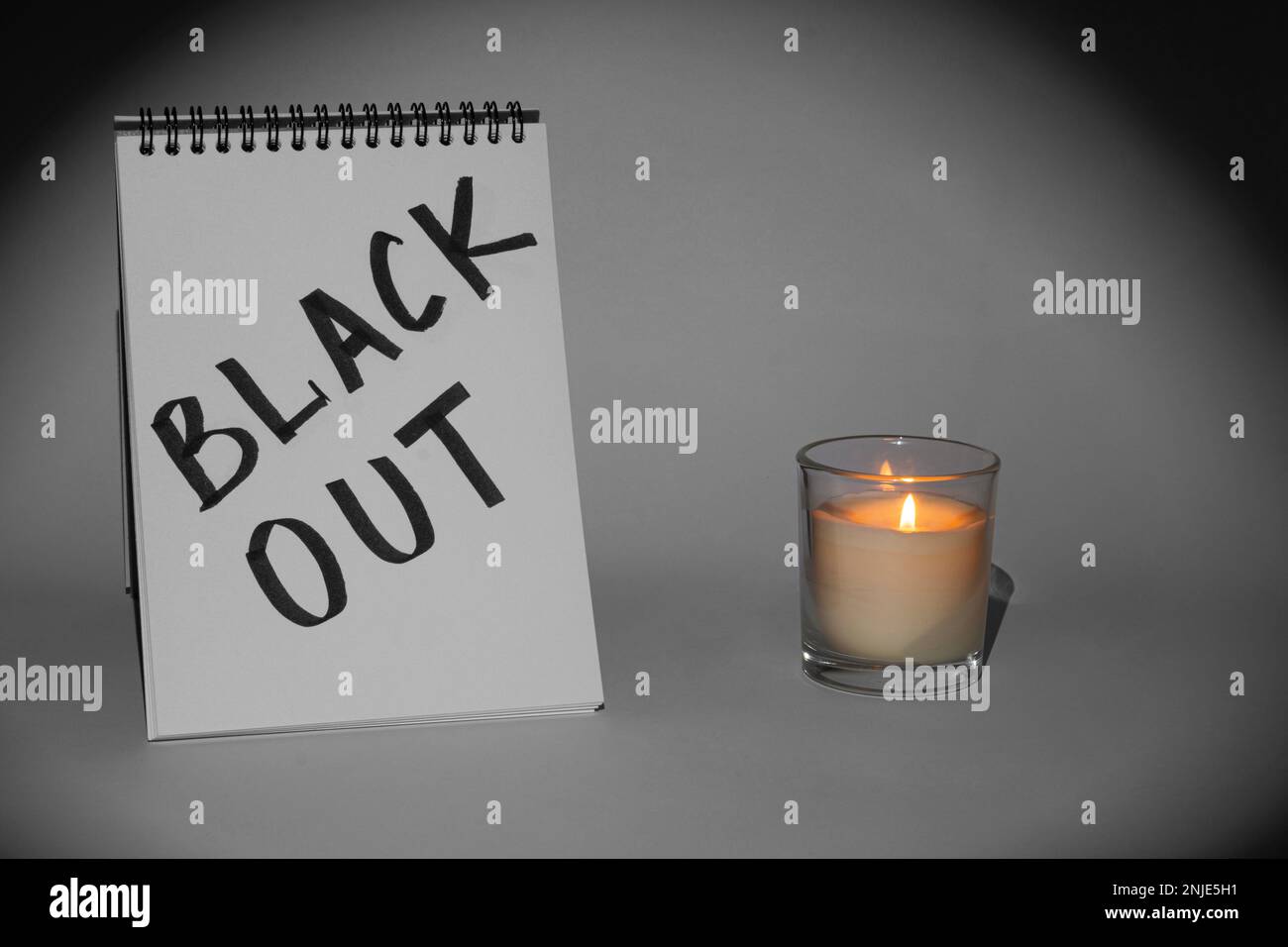 Notepad with the inscription 'Blackout' and a burning candle in a glass on a gray background, illuminated by a flashlight. Monochrome picture Stock Photo