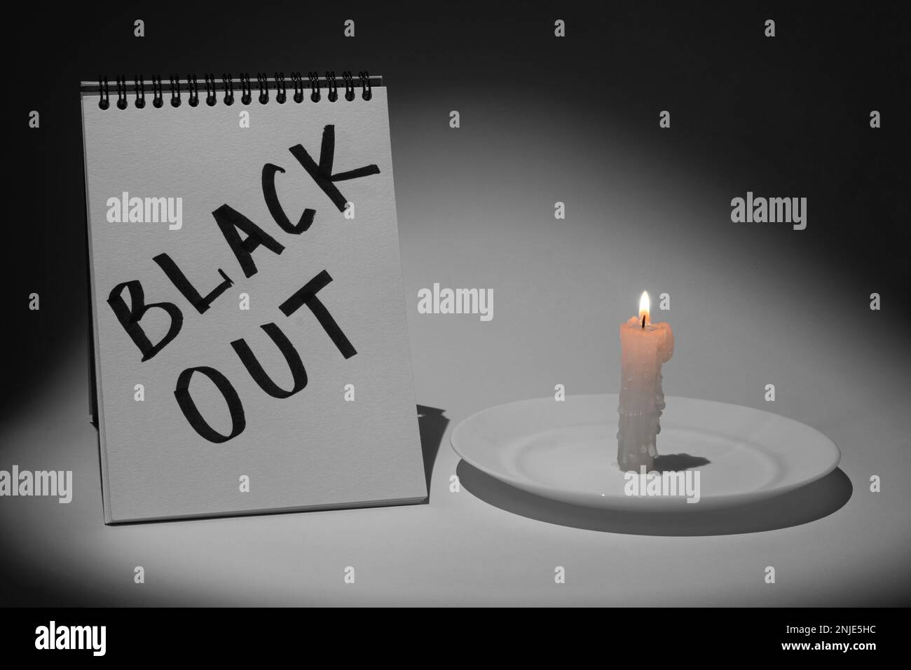 Notebook with the inscription 'Blackout' and a burning candle on a saucer on a gray background, illuminated by a flashlight. Monochrome picture Stock Photo