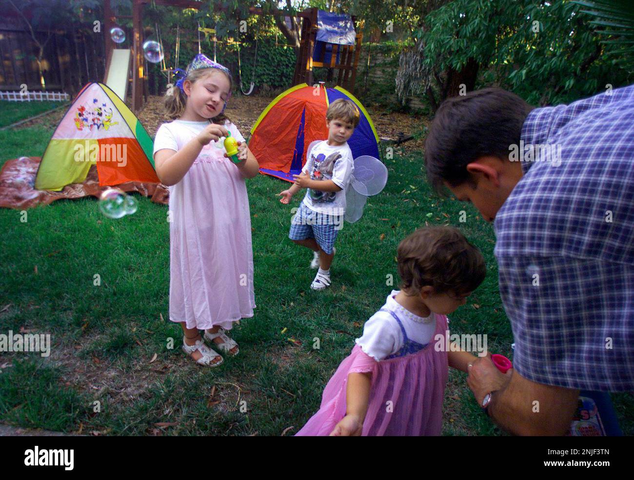 CCFRINITE01d3-C-25AUG00-CF-LH--Emily Sauter (right), 3 years old, with her dad Andy, who is helping her get ready to blow bubbles. Her sister, Sarah Sauter (left), 7 years old, is blowing bubbles, while Tyler Sanberg (middle), 4 years old, plays in the backyard. BY LIZ HAFALIA/THE SAN FRANCISCO CHRONICLE (LIZ HAFALIA/San Francisco Chronicle via AP) Stock Photo