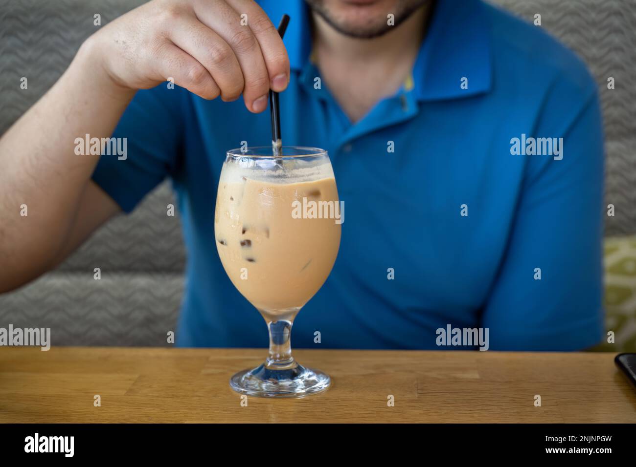 Hand of a man stirring iced latte coffee with a straw on a wooden table. Cold brew refreshment summer drink. Stock Photo