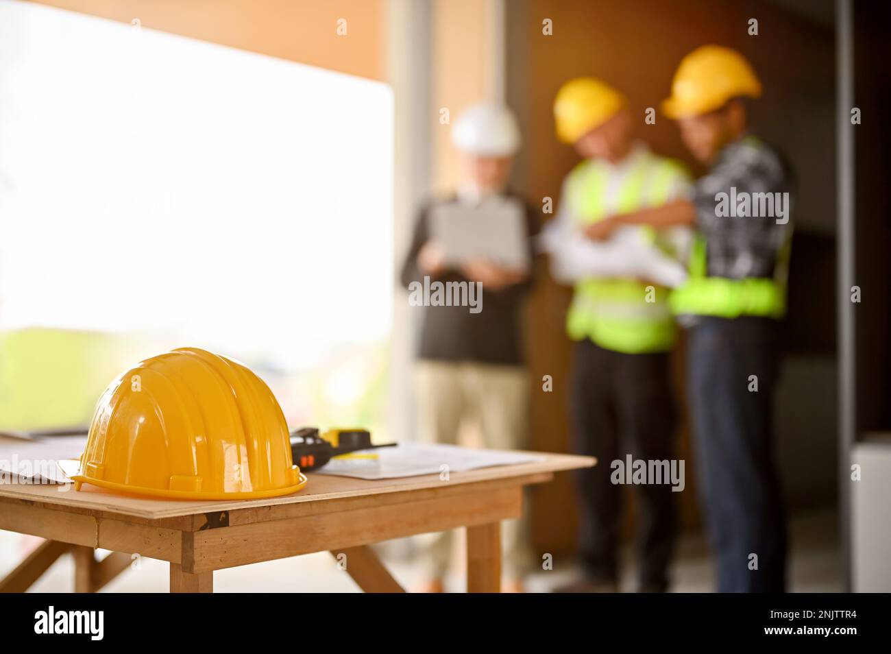 A yellow hardhat, a walkie-talkie, a measuring tape and a blueprint on a table over blurred background of a group of construction workers discussing w Stock Photo