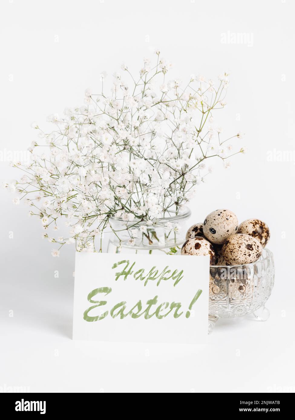 White gypsophila in glass vase, white sheet with words Happy Easter, quail eggs in crystal bowl on light backdrop. Festive creative monochrome image. Stock Photo