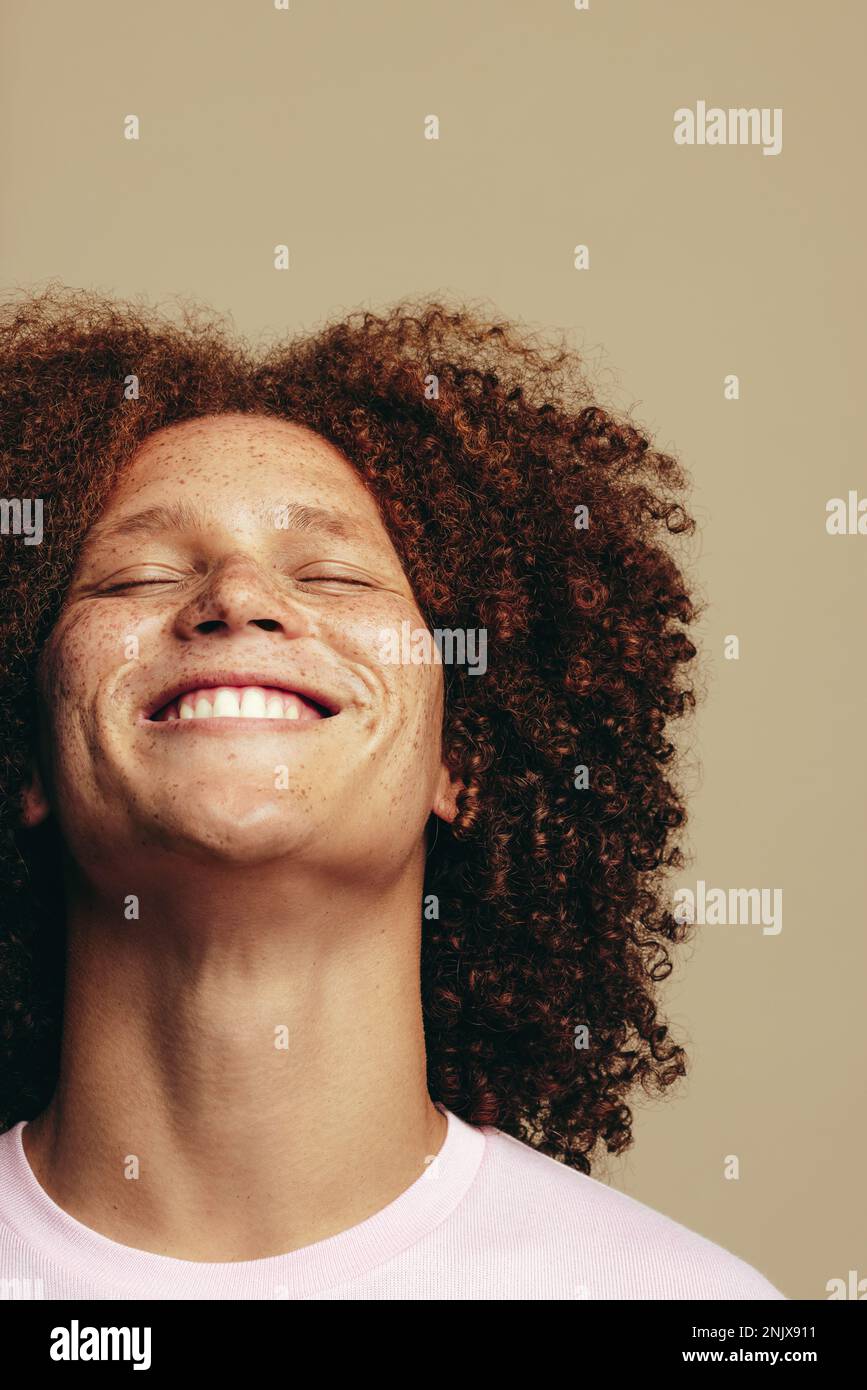 Young man with freckles and curly ginger hair smiles with his eyes closed in a studio, radiating self-love and self-confidence. Happy young man embrac Stock Photo