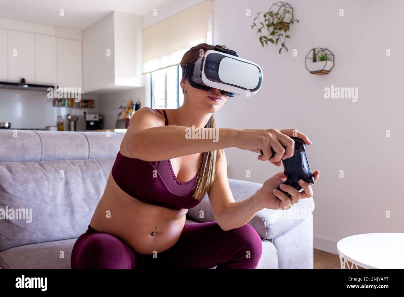 Concentrated pregnant female in sportswear and VR headset playing videogame  with joystick while sitting on sofa in living room Stock Photo - Alamy