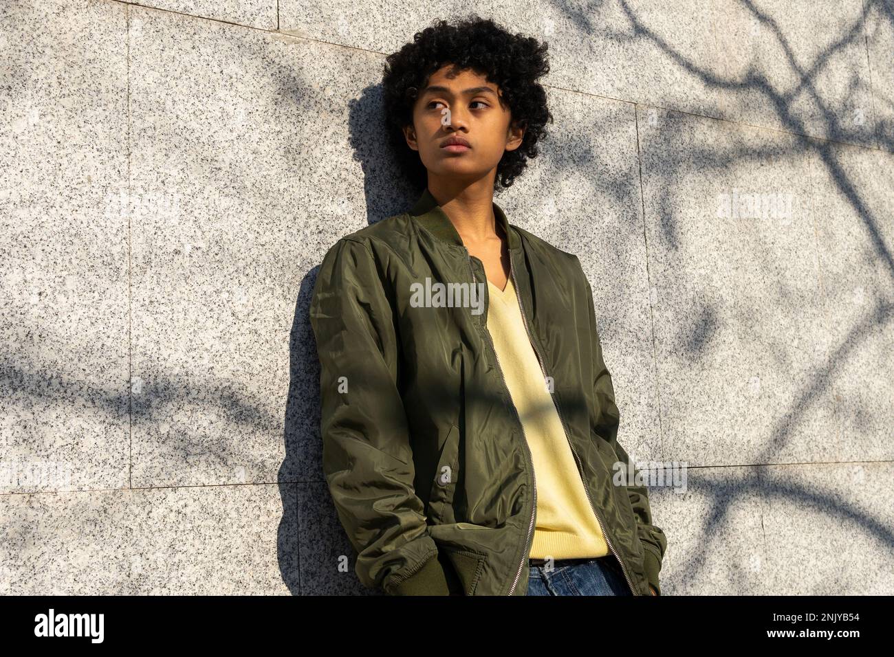Confident young Asian male with Afro hairstyle in yellow sweatshirt looking away against tiled wall with shadow in sunlight Stock Photo