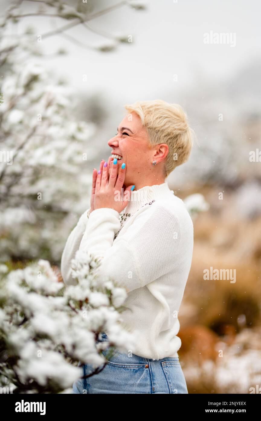 Side view of happy adult blond haired female in warm sweater smiling and looking away near tree branch during winter holidays Stock Photo