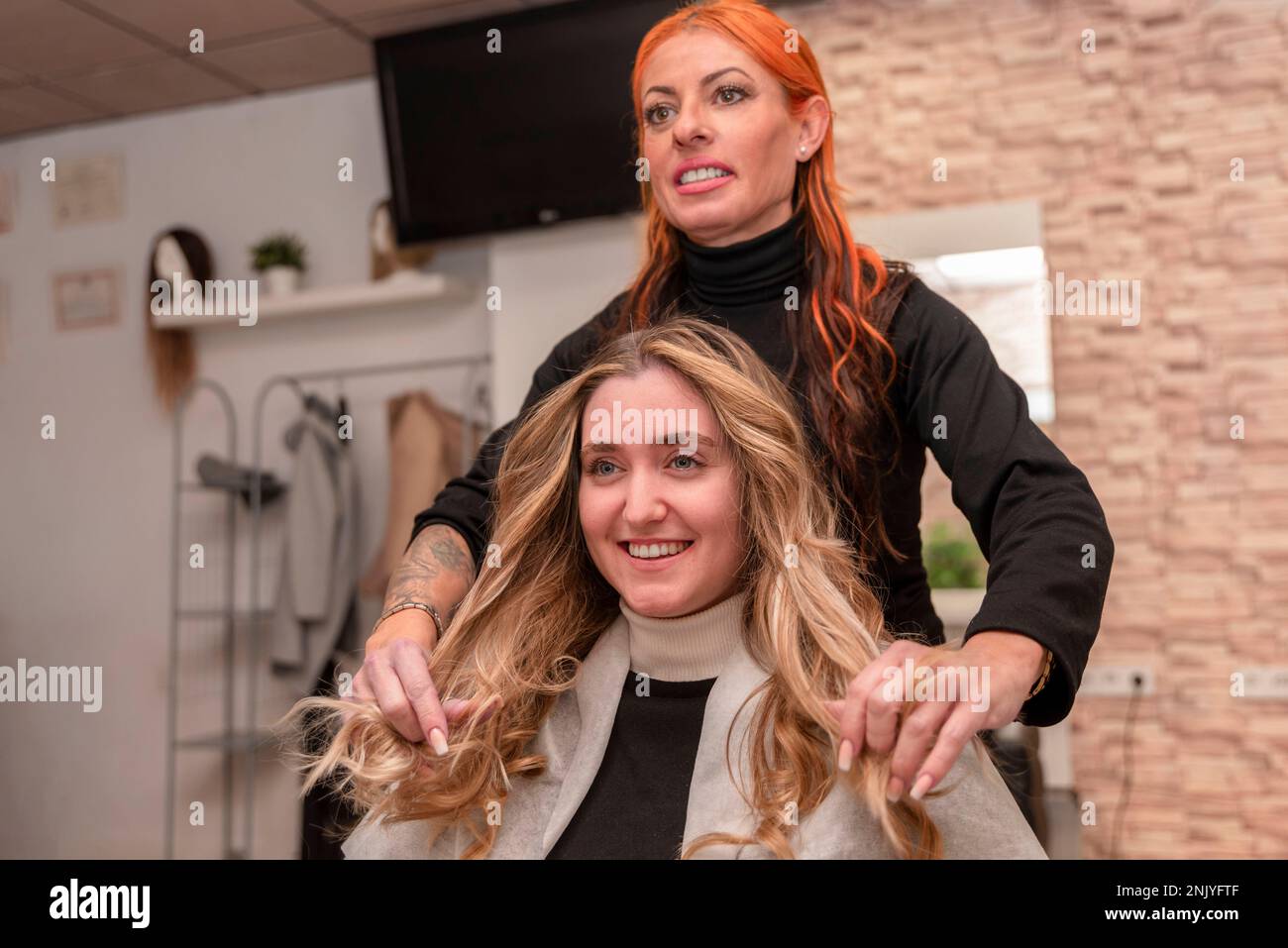 Cheerful female hairdresser smiling and checking hairstyle of happy young blond haired client in beauty salon Stock Photo