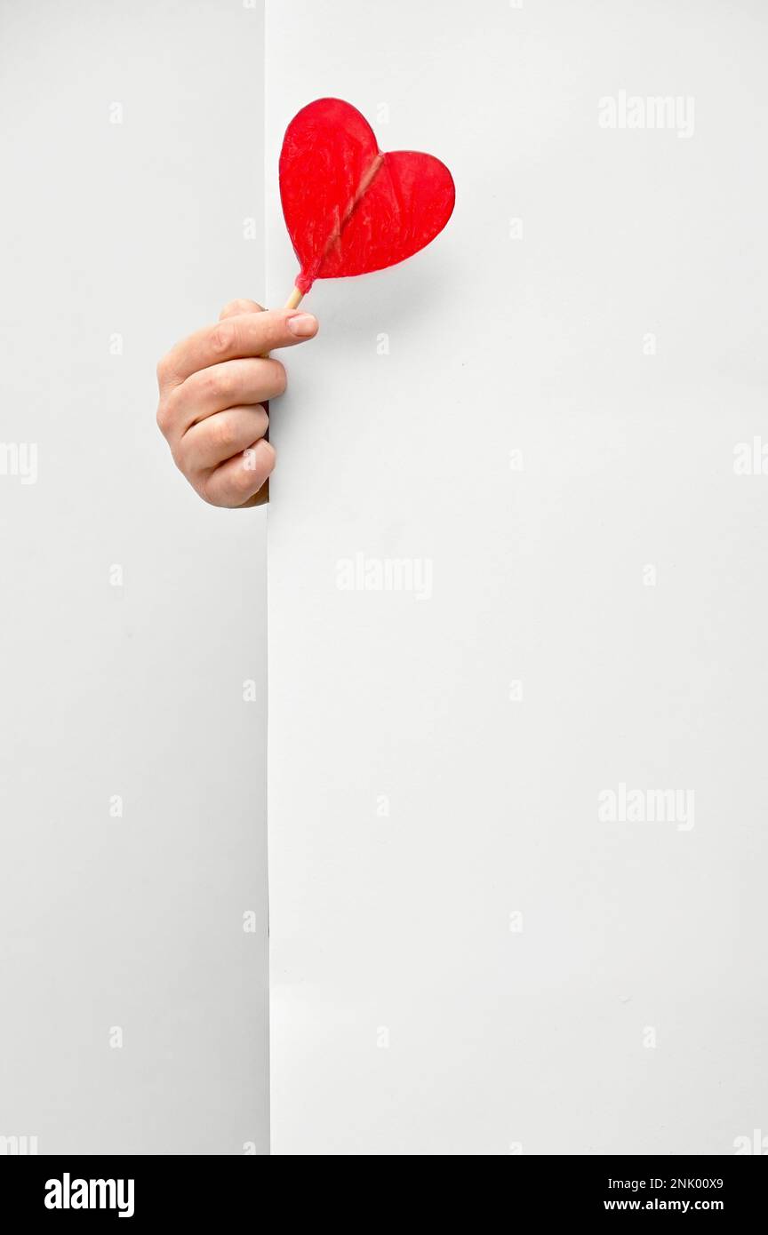Male hand holding and offering Heart Shaped Lollipop behind white wall background Stock Photo