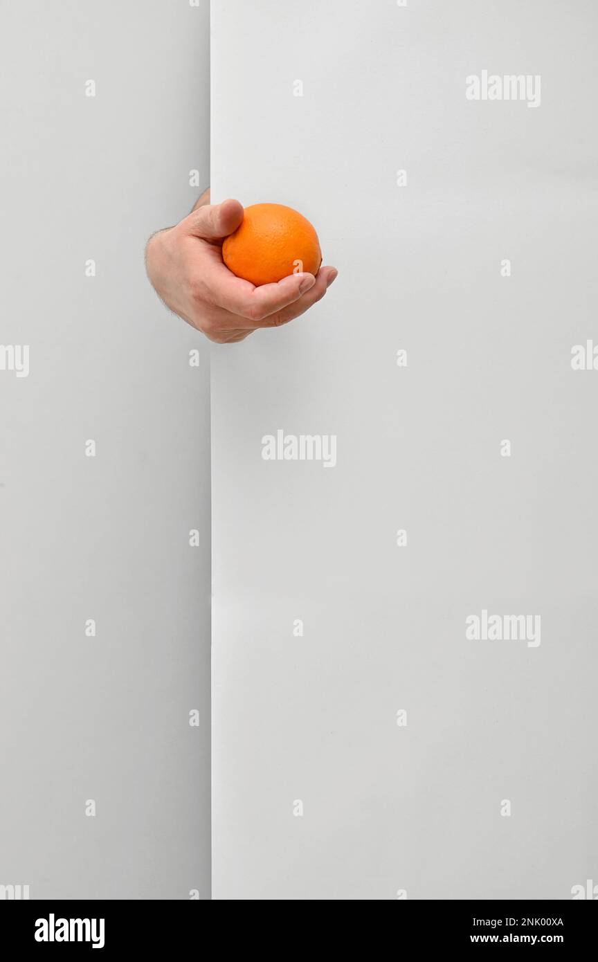 Male nutritionist hand holding and offering orange fruit behind white wall background Stock Photo
