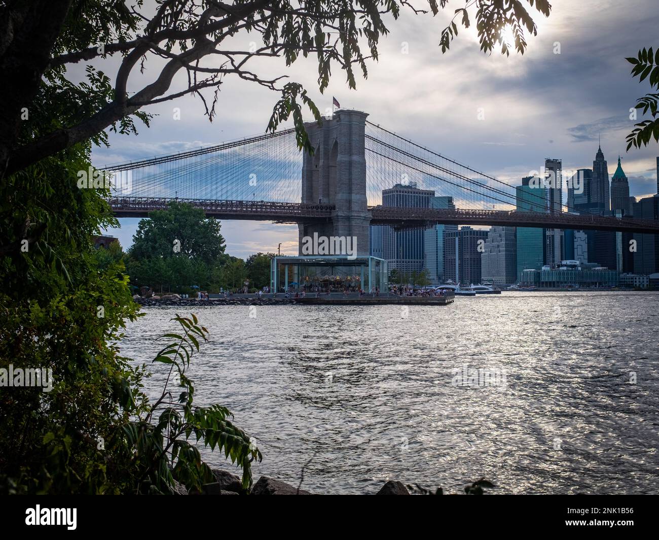 New York City, United States - August 22, 2022: Foliage framed view of the Brooklyn bridge seen from Brooklyn with unrecognizable people from afar. Stock Photo