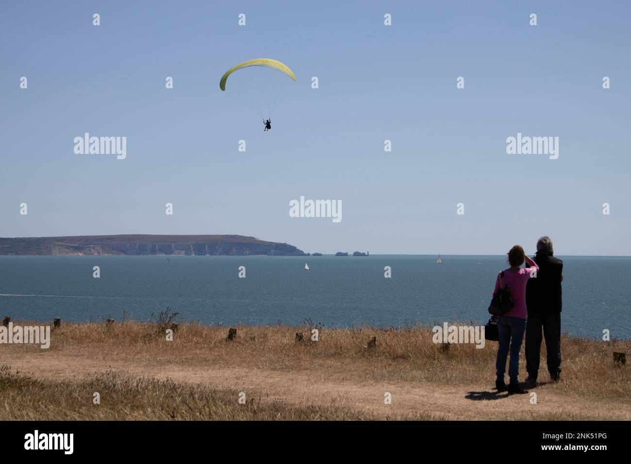 A couple watch a paraglider in the air at Barton-On-Sea, South Coast of England with the Isle of Wight in the distance, United Kingdom Stock Photo