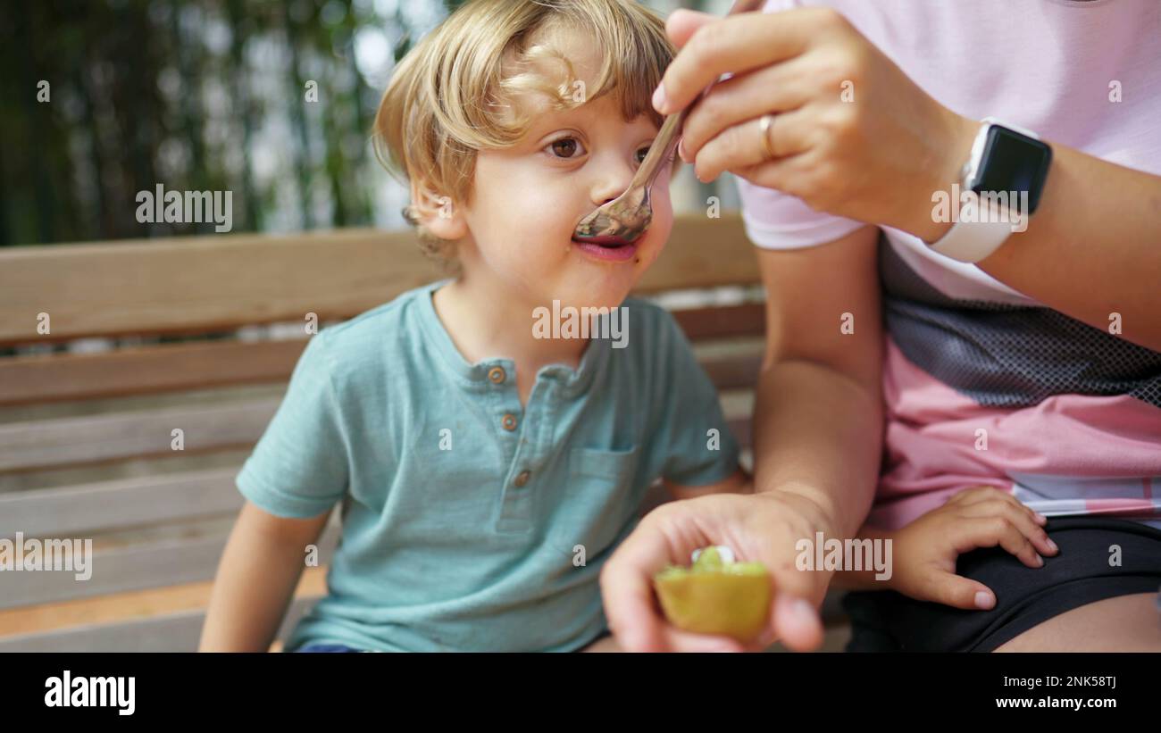 Child snacking kiwi fruit outside sitting on park bench. Closeup mother ...